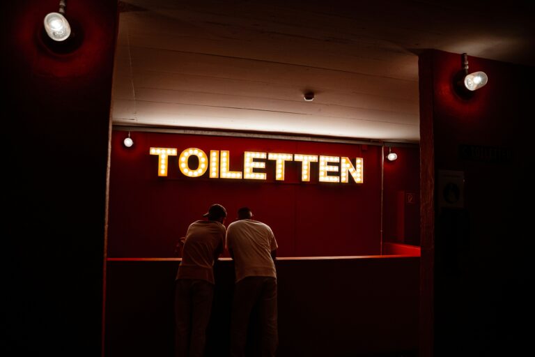 A couple of men standing in front of a red sign with white text - How to Replace a Toilet Flapper to Stop Running Water