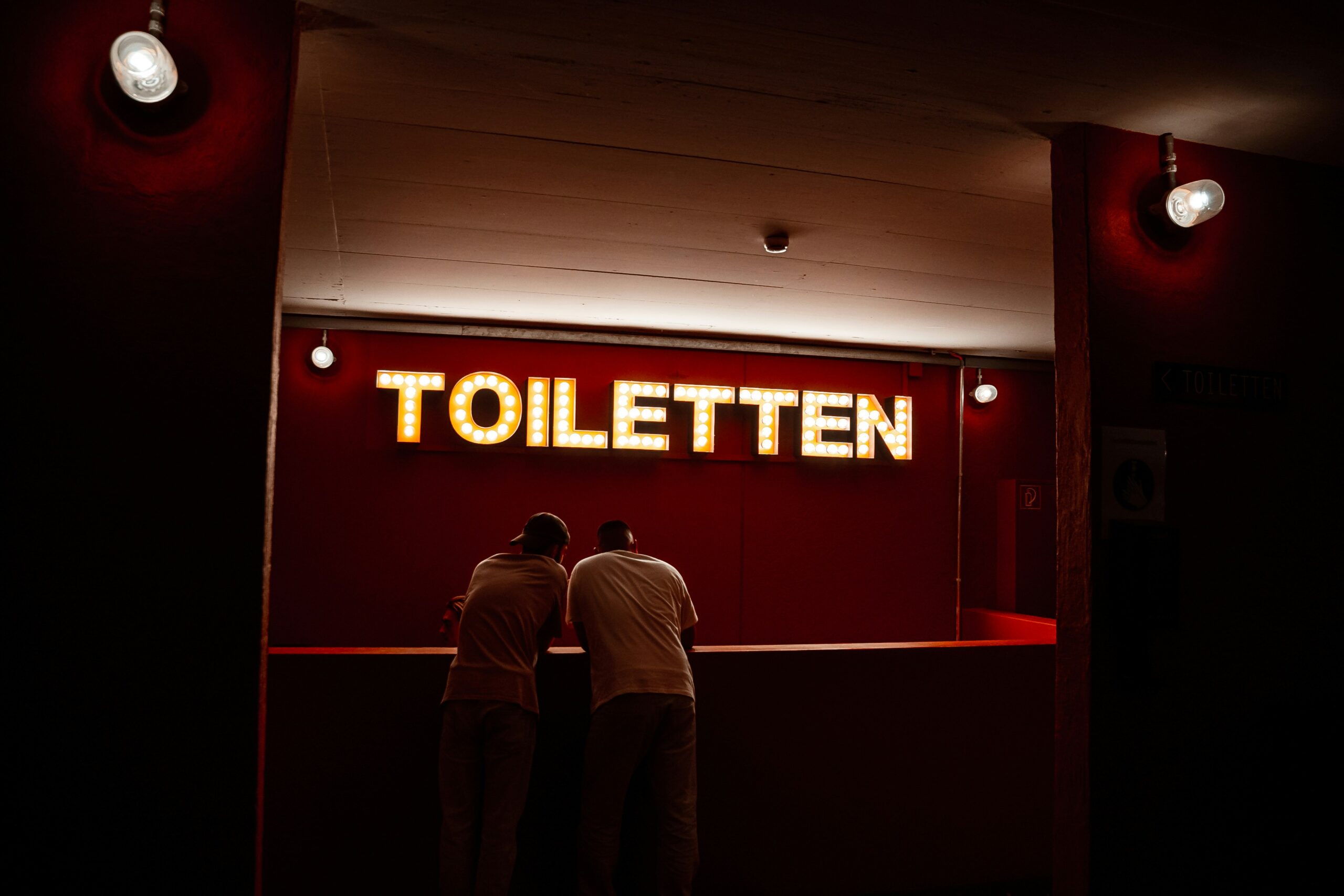 A couple of men standing in front of a red sign with white text - How to Replace a Toilet Flapper to Stop Running Water